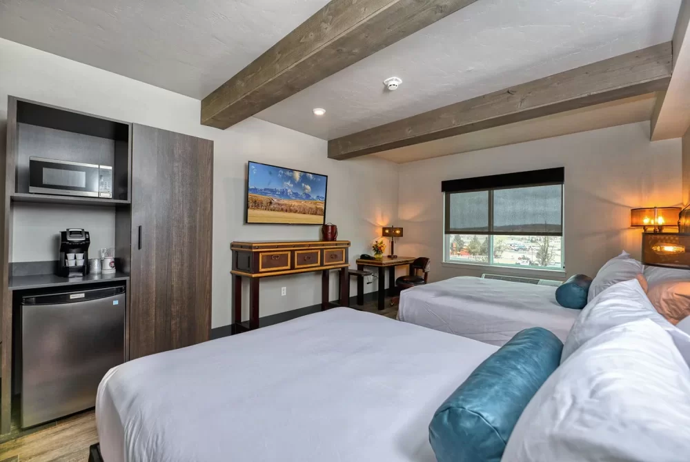 Spacious hotel room with two beds, a desk, a TV, a cabinet, and a window with a view—perfect for guests searching for Hotels in Madras OR. The kitchenette area features a fridge, microwave, and coffee maker for your convenience. Exposed wood beams on the ceiling add a rustic touch to this comfortable and inviting space.