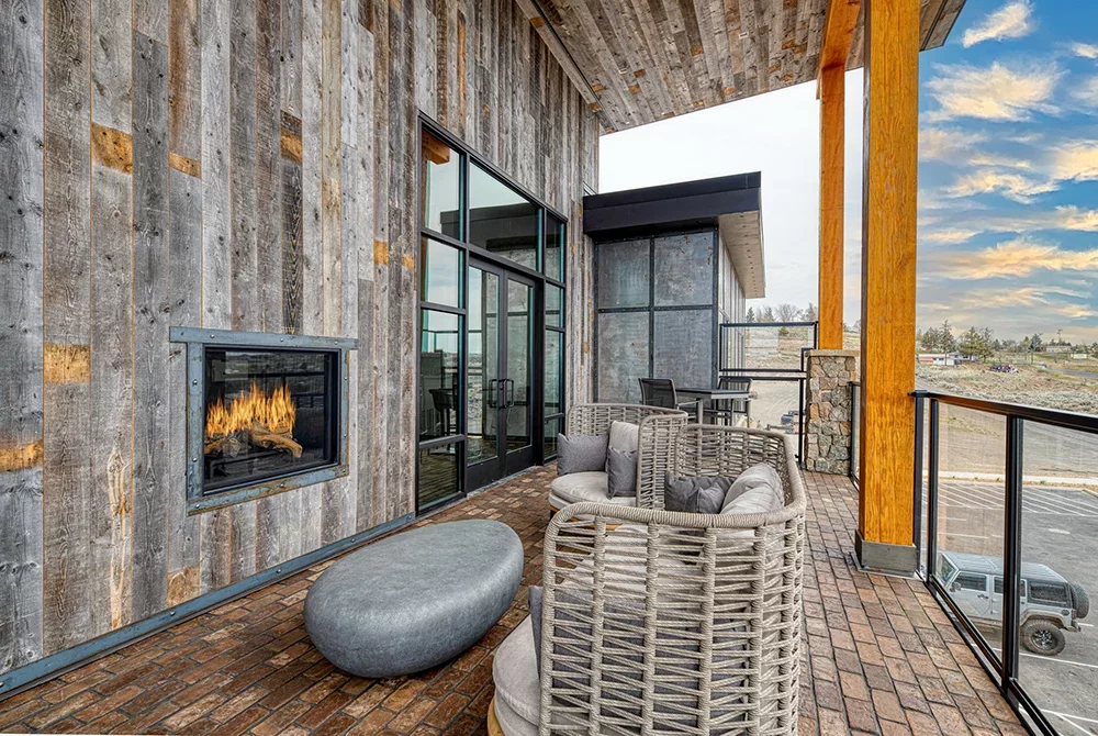 Modern patio with wicker chairs, a stone ottoman, a wall-mounted fireplace, and glass doors, set against rustic wood walls and a scenic outdoor view under a partly cloudy sky—an inviting ambiance reminiscent of premier hotels in Madras OR.
