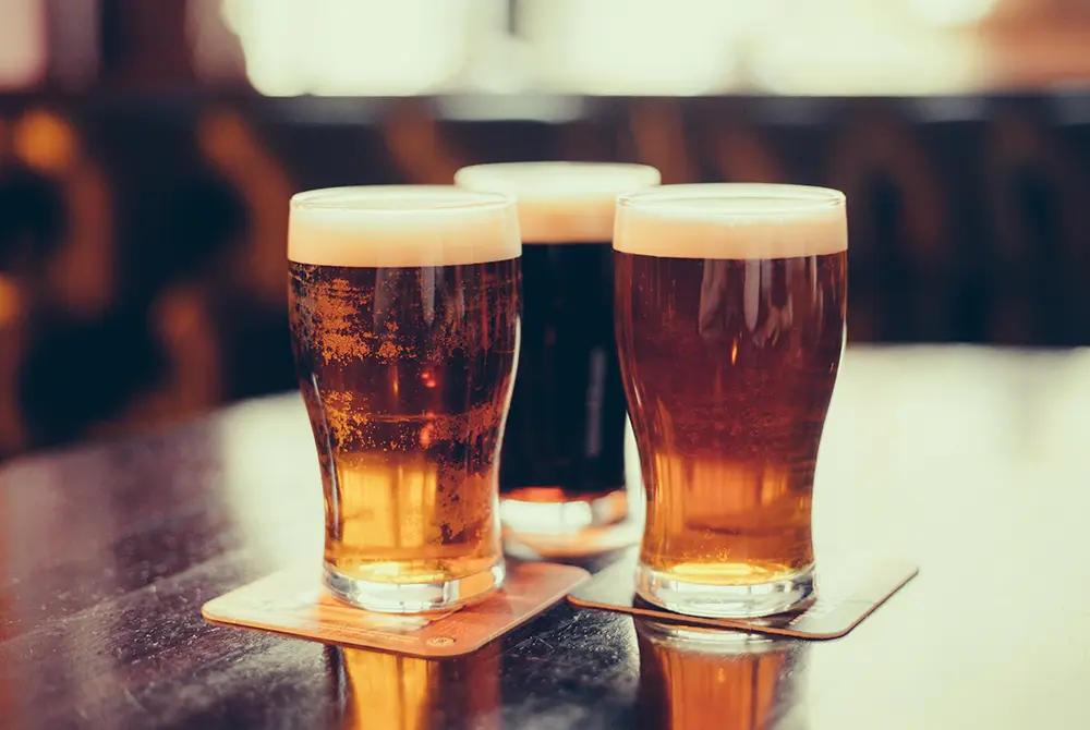 Three pint glasses of beer, each with a frothy head, sit on coasters atop a wooden table. The beers are different colors: pale, amber, and dark. The background is softly blurred—a perfect snapshot of the local craft scene and one of the best things to do in Madras OR for beer enthusiasts.