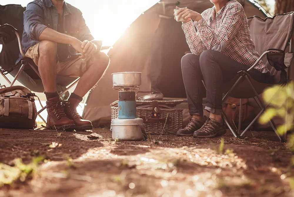 Two people sit in camping chairs near a tent, with a small stove and cooking pots between them. Both are casually dressed, enjoying a drink outdoors in a sunlit, natural setting—just one of the relaxing things to do in Madras OR for those who love connecting with nature.