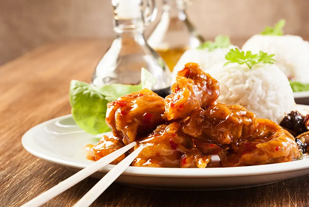 Chopsticks picking up a piece of saucy meat on a plate, with rice garnished with cilantro and sauce on the side—a delicious meal that could easily top your list of things to do in Madras OR. Glass bottles in the background rest on a wooden table, enhancing the inviting atmosphere.