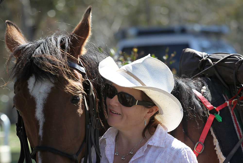 A woman in a white hat and sunglasses smiles while standing beside a brown horse with a white stripe on its face. The horse is wearing a bridle and saddle—perfect for those looking for unique things to do in Madras OR.