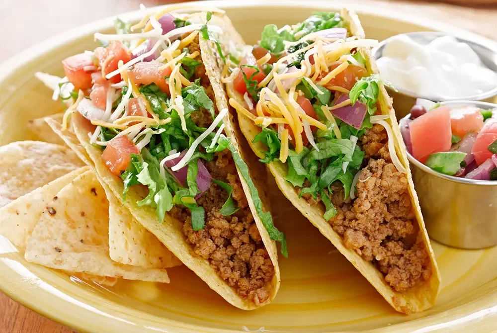 Two crunchy beef tacos topped with shredded cheese, lettuce, tomatoes, and onions are served with crispy tortilla chips, a side of salsa, and sour cream on a bright yellow plate—the perfect meal before exploring all the exciting things to do in Madras OR.